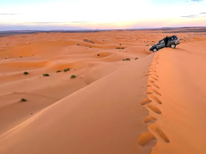 A vehicle traverses the vast Sahara Desert, surrounded by endless sand dunes under a clear blue sky Erg Chebbi Dunes 