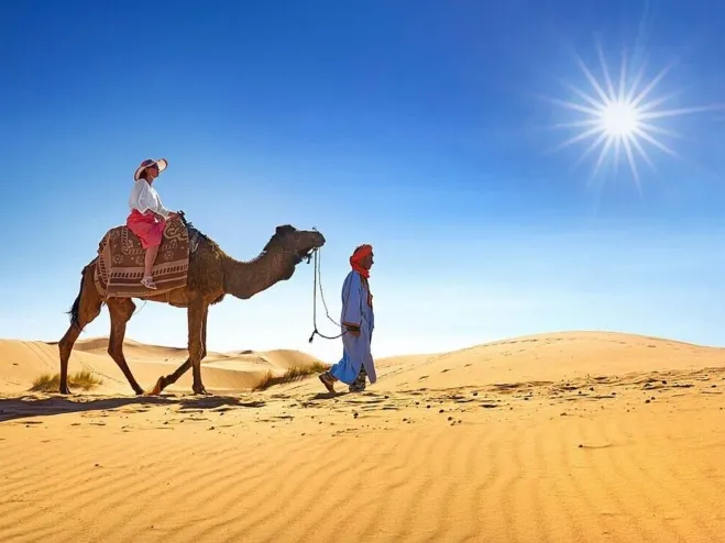 Two individuals riding a camel across a vast desert landscape under a clear blue sky moroccan desert tour