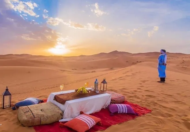 A serene sunset dinner set against the backdrop of the Sahara Desert, featuring elegant table settings and warm hues Marrakech desert excursions