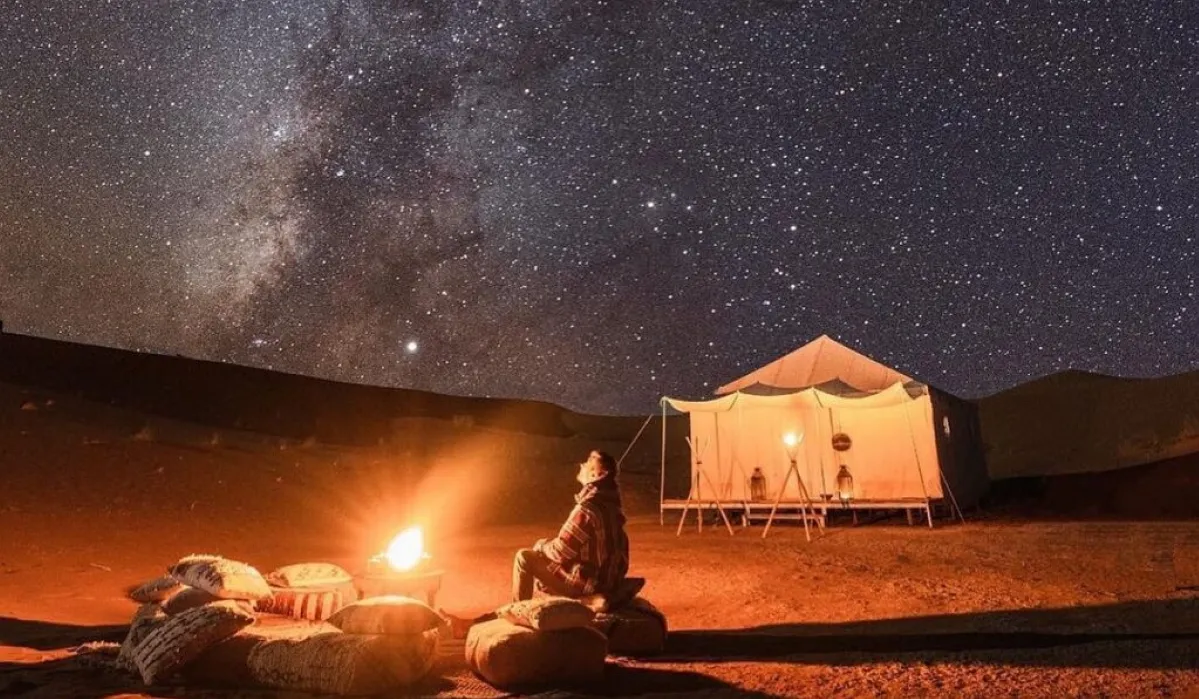 A tent in the desert under a starry sky, with a warm fire glowing nearby, creating a serene and inviting atmosphere Morocco desert night