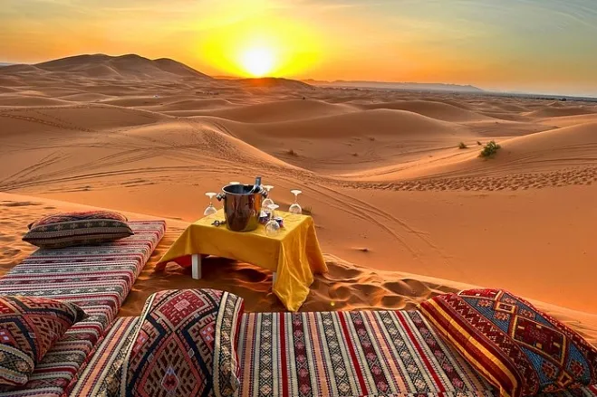 A table and chairs arranged in the desert, illuminated by the warm hues of a stunning sunset morocco desert tour