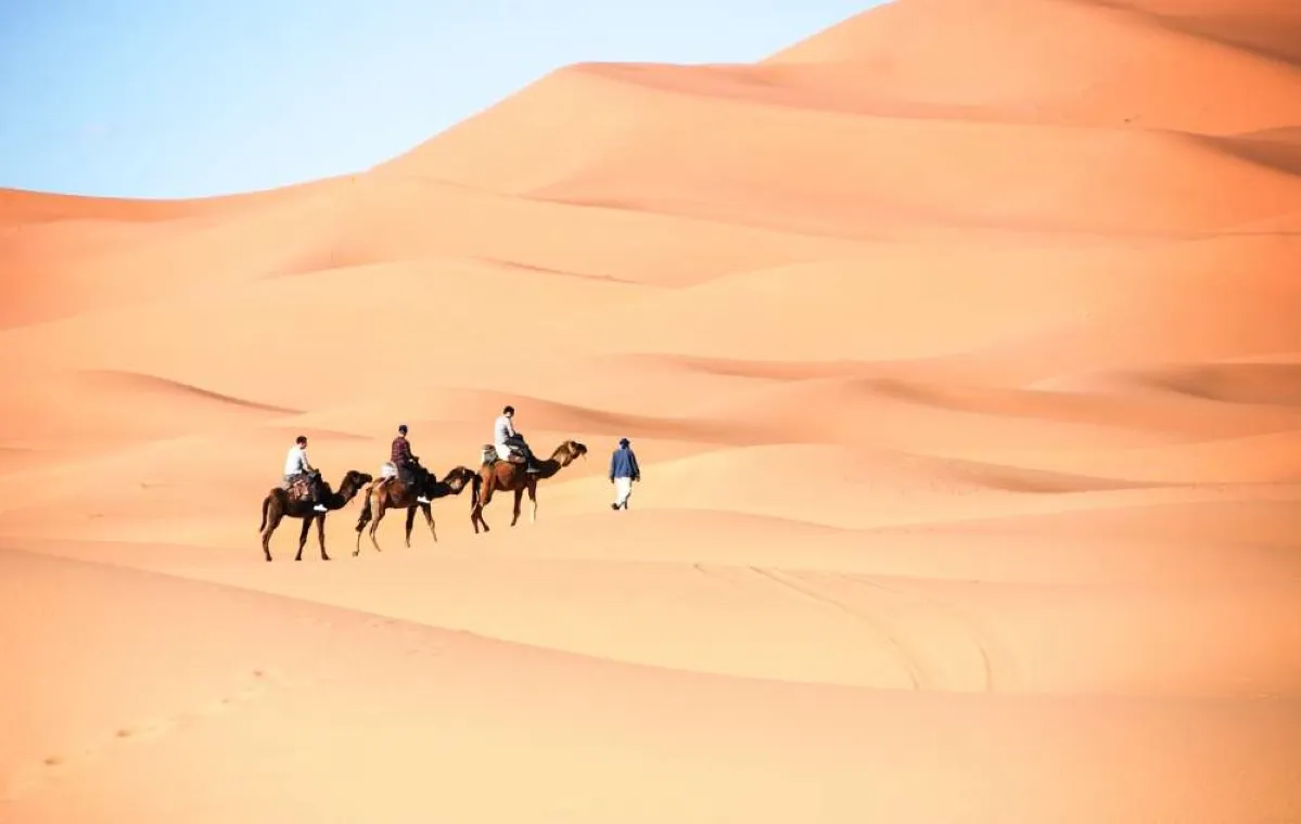 Three individuals riding camels traverse a vast desert landscape under a clear sky Marrakech desert excursions