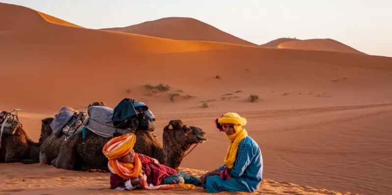 Two individuals in vibrant attire seated on the sand, surrounded by camels in the Moroccan desert landscape.