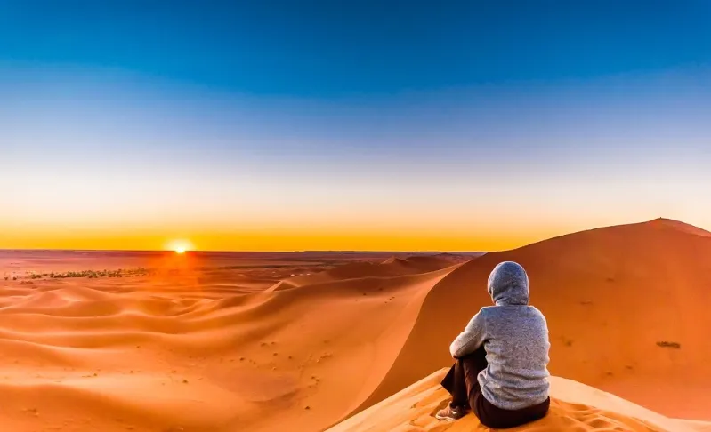 A Woman sits on the sand dunes of the Sahara Desert at sunset, capturing the serene beauty of Morocco's landscape.