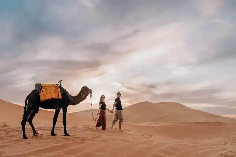  A couple strolls through the Moroccan desert, accompanied by a camel, enjoying a family tour experience. 