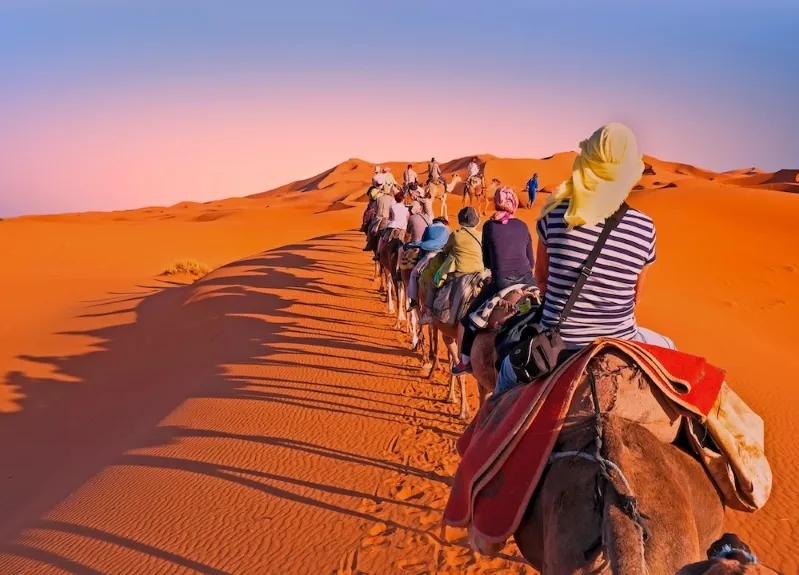 Tourists riding camels across the vast Sahara desert during a journey from Fes, showcasing the stunning landscape. 