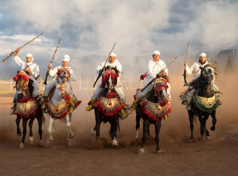 Four men in white outfits riding horses through dirt at a vibrant Moroccan festival.