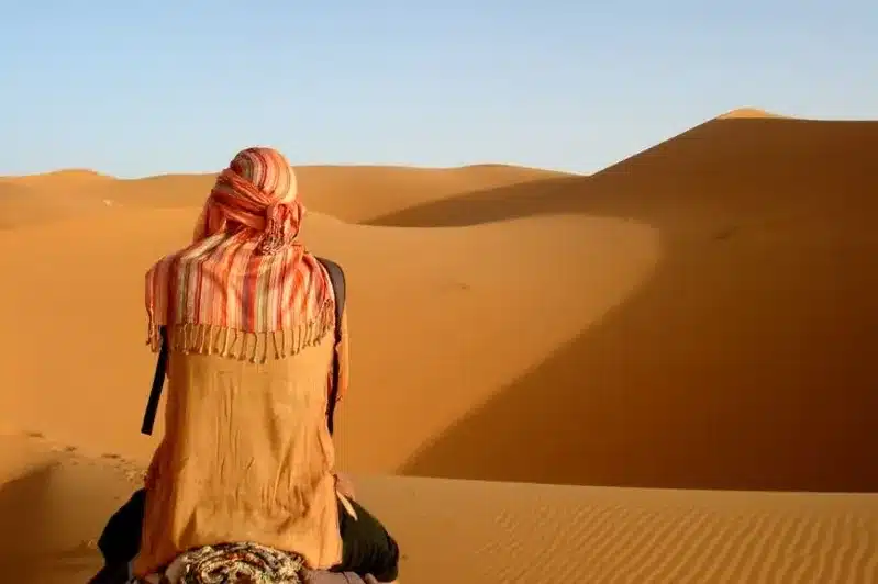 A person wearing a scarf sits atop a camel, surrounded by the vast expanse of the Sahara Desert in Morocco.