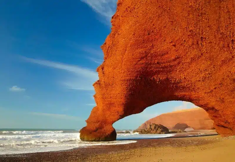 The red rock arch stands majestically at a desert beach, highlighting the beauty of beach resorts in morocco.

