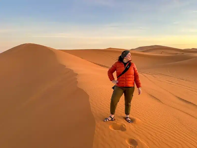 A woman stands confidently in the vast expanse of the Sahara Desert, surrounded by golden sand dunes in Morocco.