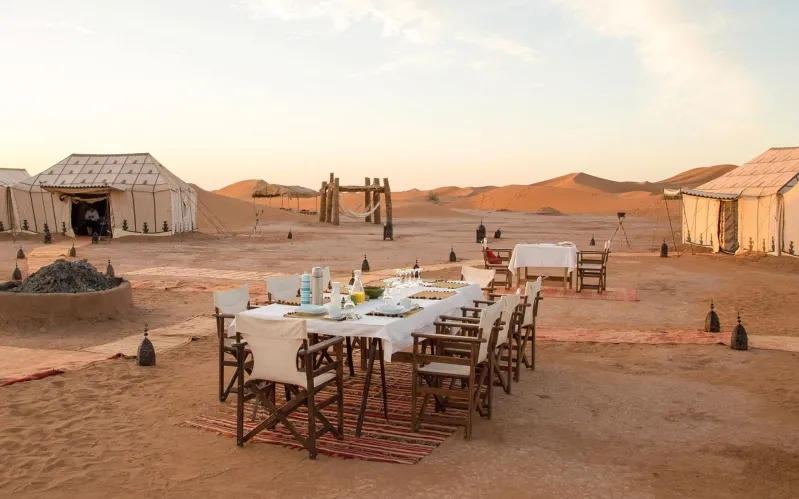 A table elegantly arranged in the Moroccan desert, surrounded by tents and chairs, showcasing a serene outdoor setting.