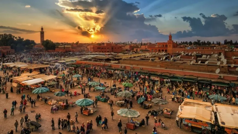 A vibrant sunset casts warm hues over the bustling Marrakech market, framed by historical sites in the background. 