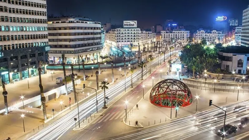 A bustling Gueliz street in Marrakesh at night, illuminated by traffic and surrounded by vibrant buildings.
