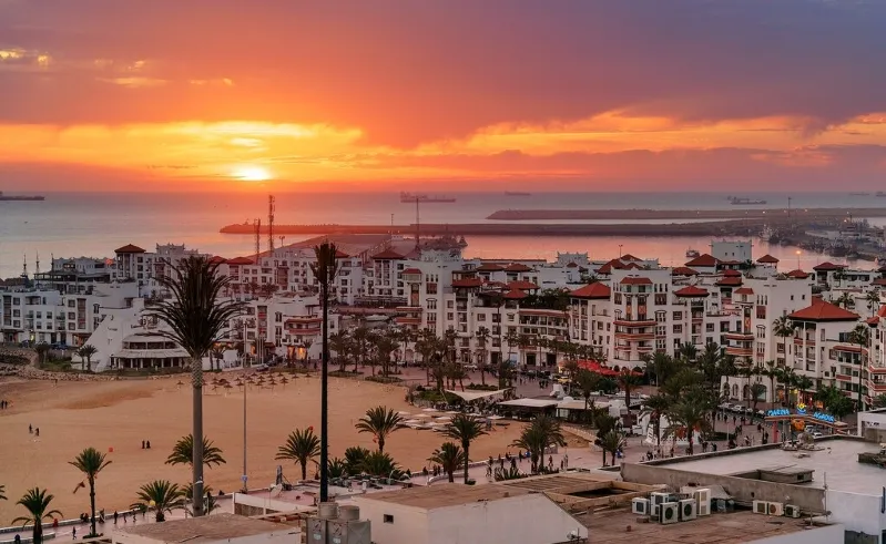 A stunning sunset casts a warm glow over the city of Marrakech, Morocco, with silhouettes of buildings against the vibrant sky.
