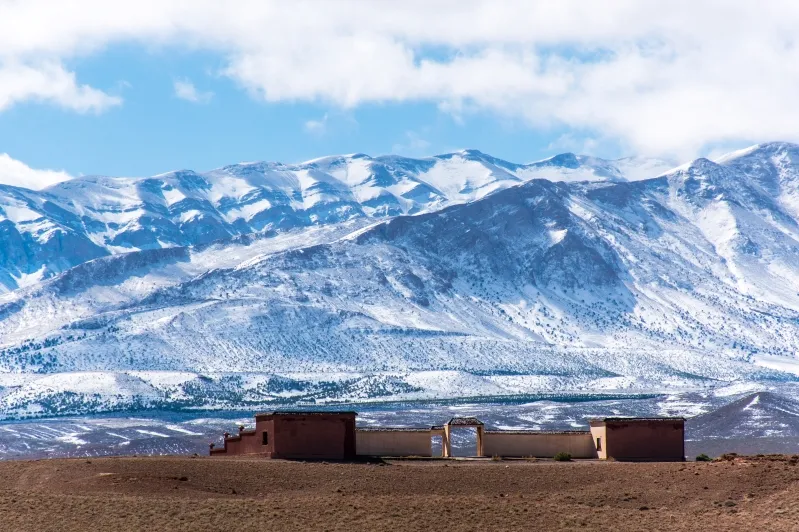 A building stands alone in the desert, with the snow-capped Atlas Mountains range majestically behind it.