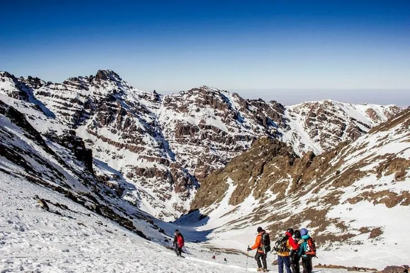 A group of hikers ascends a snowy mountain, surrounded by snow-covered peaks under a clear blue sky.