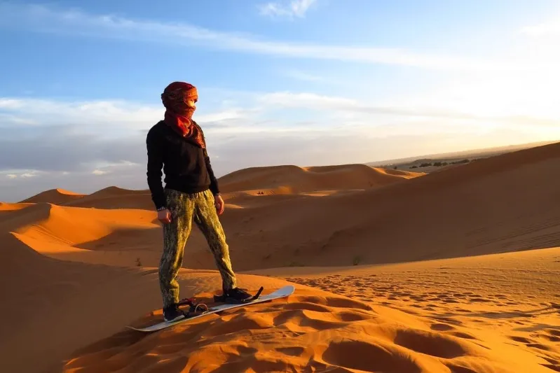 A person stands on a snowboard in a desert landscape, with a camel ride in the background near Casablanca.