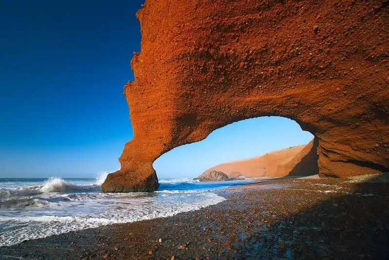 A red rock arch on a beach in Morocco, with waves crashing against it, showcasing the natural beauty of the coastline. 