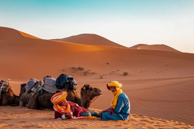 Two people dressed in colorful clothing relax on the sand with camels, capturing the essence of yoga in Marrakech.