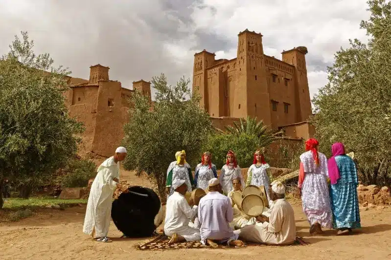 A group of individuals in traditional attire joyfully playing drums, celebrating with almond ghriba treats nearby. 