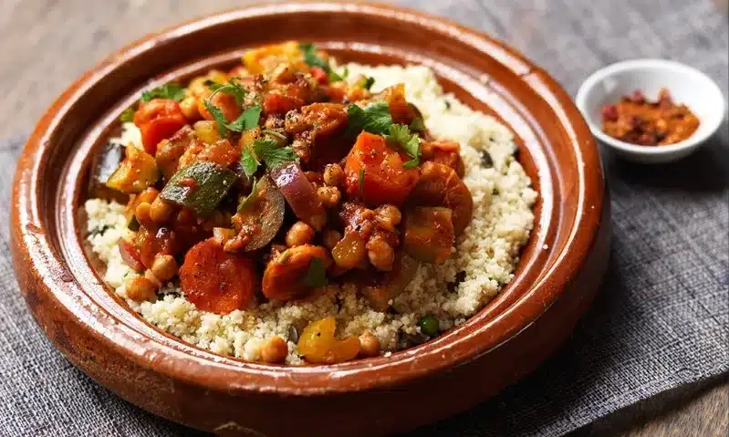 A vibrant plate of Moroccan couscous topped with colorful vegetables, accompanied by almond ghriba cookies on the side.