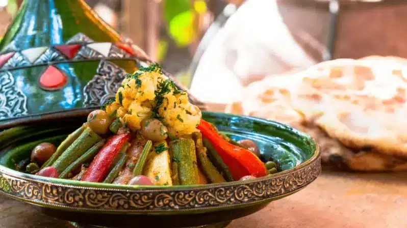 A green bowl filled with vegetables and bread sits on a table, accompanied by a serving of the beef Moroccan tagine.
