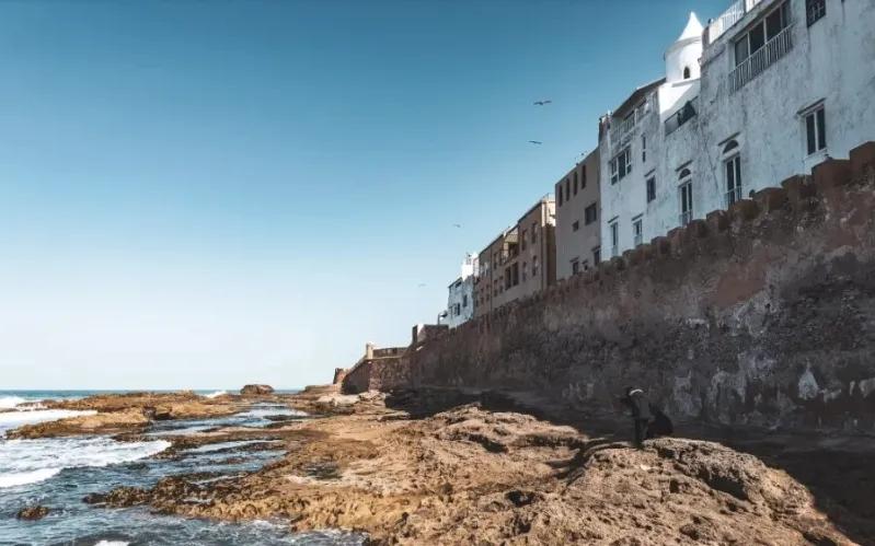 A man walks on a rocky beach in Morocco, with the ocean waves nearby, highlighting the Morocco travel advisory.