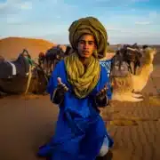 A man wearing a blue turban sits in the desert, representing the Berber culture in Morocco.