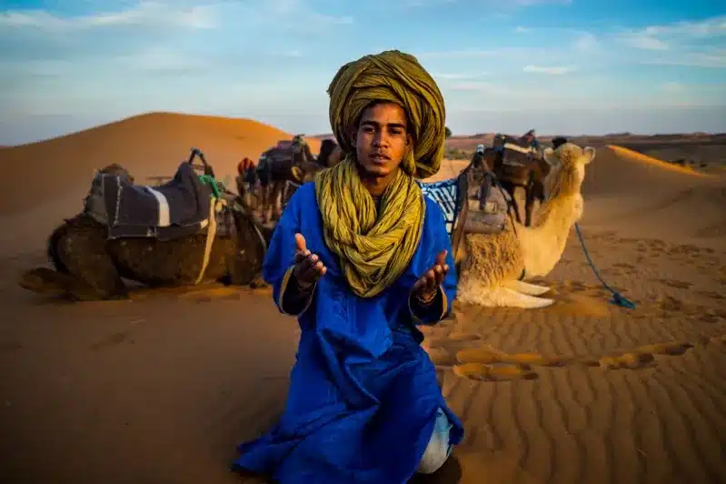 A man wearing a blue turban sits in the desert, representing the Berber culture in Morocco.