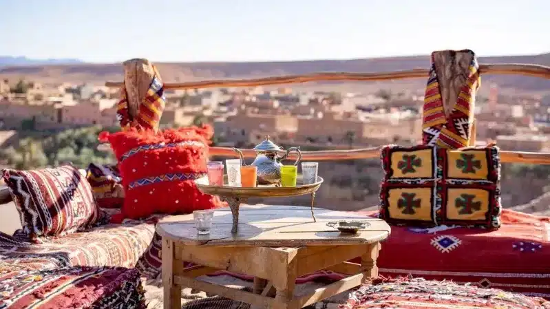 A table adorned with pillows and a rug, set against a stunning mountain backdrop, promoting Moroccan travel experiences.
