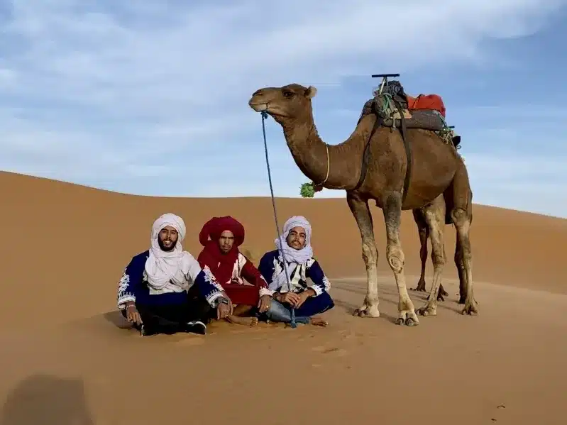 Three individuals in traditional Moroccan attire sit on the sand beside a camel, showcasing a cultural experience in Morocco.