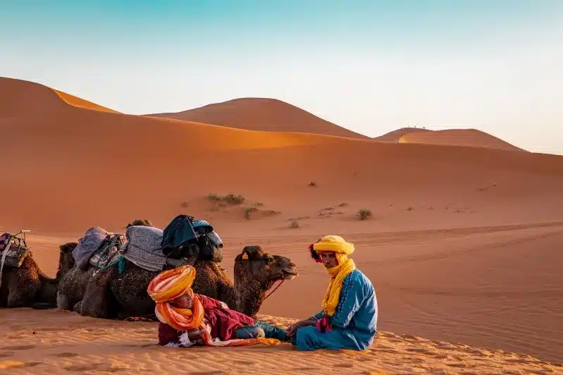 Two individuals in bright clothing relax on the sand with camels, showcasing Berbers in Morocco. 