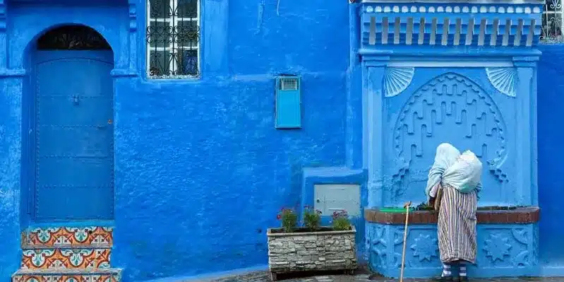 A woman in a blue dress stands confidently in front of a blue house, symbolizing safety for women in Morocco. 