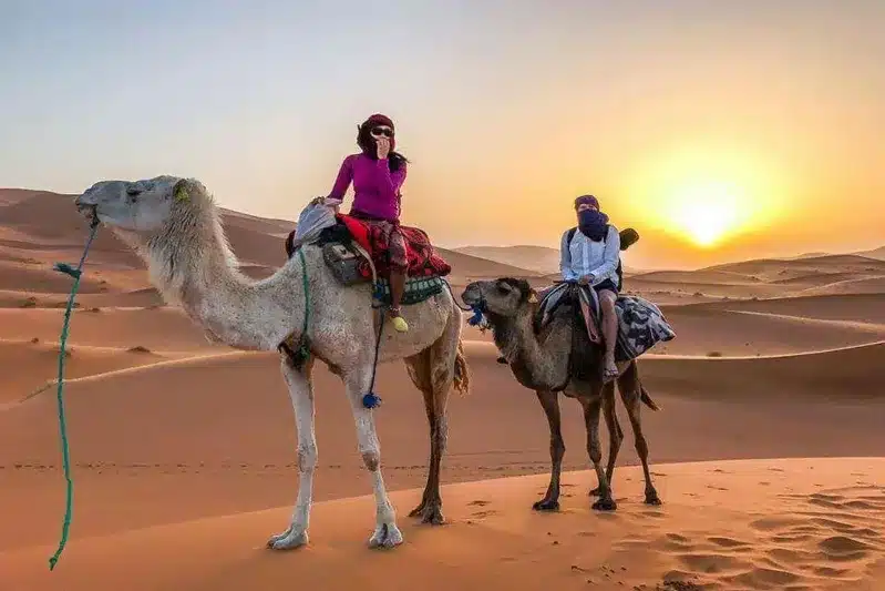 Two people riding camels in the desert during sunset, part of a Fes to Marrakech desert tour.
