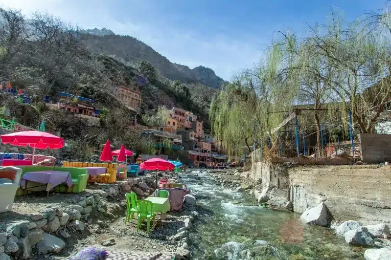 Colorful umbrellas and tables line a riverbank, creating a vibrant Moroccan dream atmosphere. 