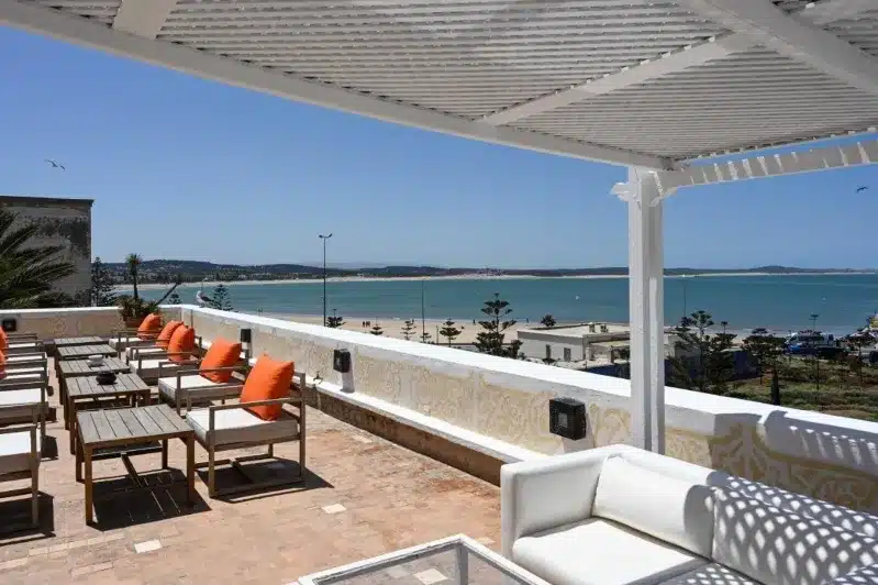 A patio with white furniture and orange cushions, offering a view of the beach, top things to do in Essaouira.