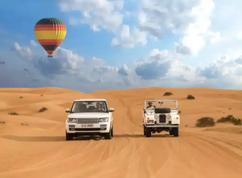 Two vehicles drive through a desert landscape, with a colorful hot air balloon floating in the background, showcasing Morocco tours.