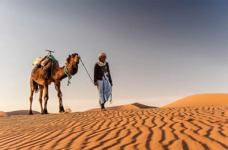  A man stands beside his camel in the Sahara Desert, showcasing the vast landscape during a Morocco tour from Marrakech.