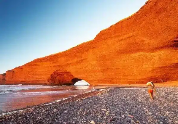 A person walks along the beach beside a large red rock during an Agafay Desert trip, with waves gently lapping at the shore.
