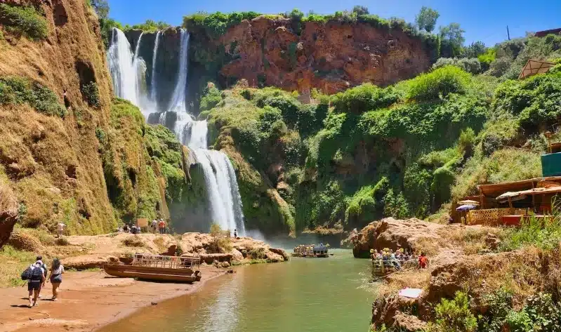 People stroll along a riverbank near a waterfall during the Ziz Valley Desert Tour.