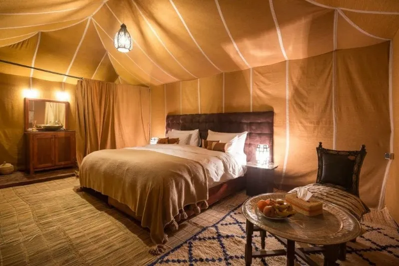 Luxurious tent interior with a cozy double bed, decorative pillows, and warm lighting. Wooden furniture and a patterned rug create a tranquil and inviting atmosphere.