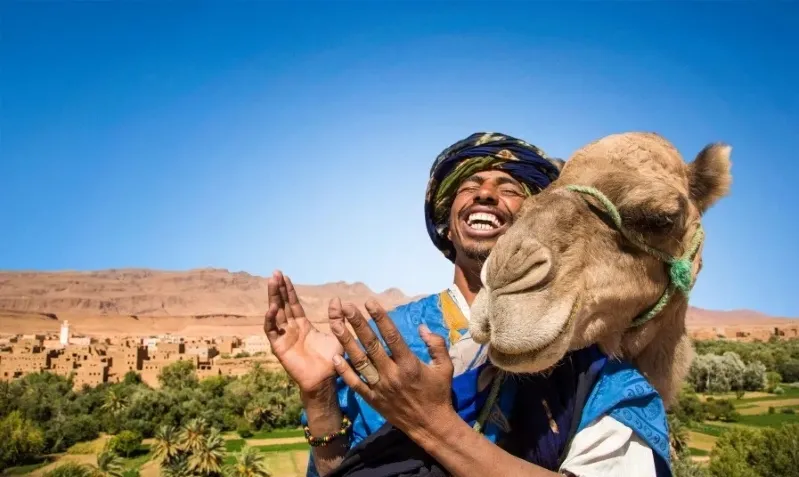 A man in a turban and blue scarf holds a camel near the Ouzoud Waterfalls, showcasing a blend of culture and nature.