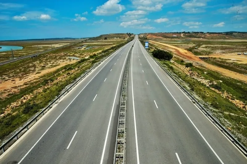 Straight empty highway stretching into the horizon on a sunny day, flanked by open grassy fields and distant hills under a blue sky with fluffy clouds. With Morocco Travel News today