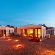Desert glamping site at dusk with three illuminated luxury tents surrounded by decorative lanterns. The sky is a deep blue, creating a serene ambiance.
