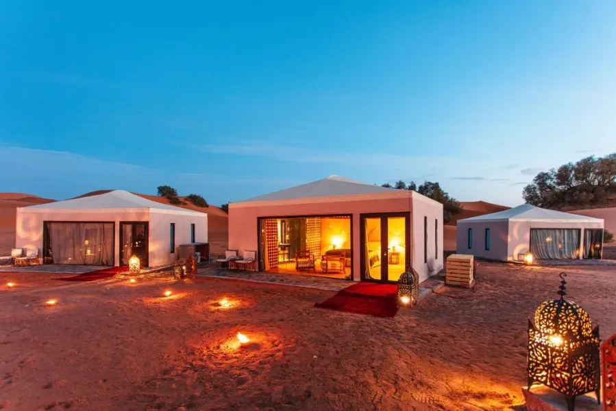 Desert glamping site at dusk with three illuminated luxury tents surrounded by decorative lanterns. The sky is a deep blue, creating a serene ambiance.