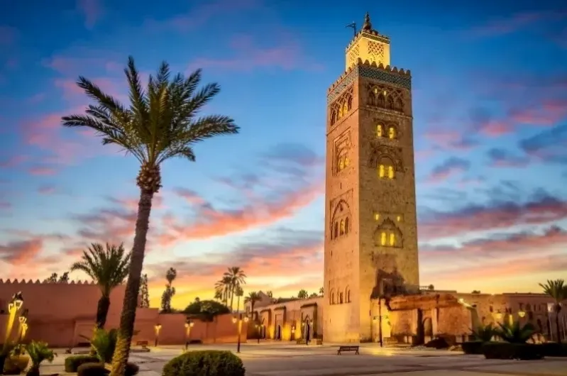 A tall, ornate mosque tower stands majestically against a vibrant sunset sky in Marrakech, surrounded by palm trees and illuminated by warm lights.
