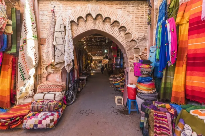 Narrow market alley with colorful textiles and rugs hanging from brick arches. Bright, vibrant colors create a lively atmosphere, inviting exploration.