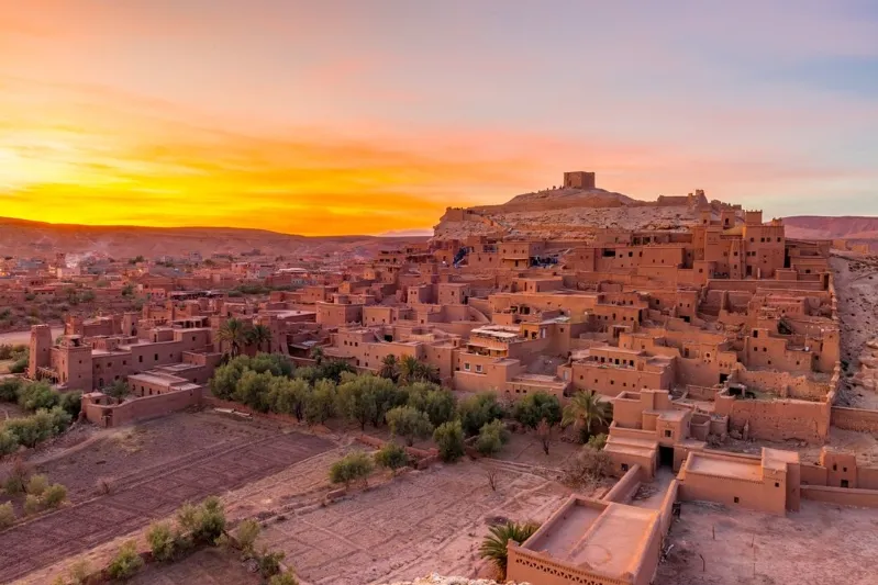 Ait Benhaddou under a vibrant sunset, with warm orange hues lighting up the sky. The ancient clay buildings and surrounding greenery create a serene, historic atmosphere.