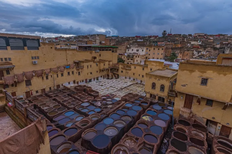 Aerial view of a traditional tannery with circular dye vats in various colors. Surrounding are beige buildings under a cloudy, dramatic sky, conveying a historic and vibrant atmosphere.Between best things to do in Fes.