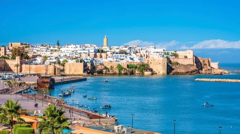 Coastal view of a historic city with white buildings and a tall tower surrounded by ancient walls. The bright blue sea and sky create a vibrant, serene scene.While our travel in Morocco.
